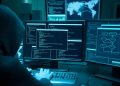 Hooded person working at a multi-monitor setup with glowing blue code and maps on the screens, creating a cybersecurity scene.