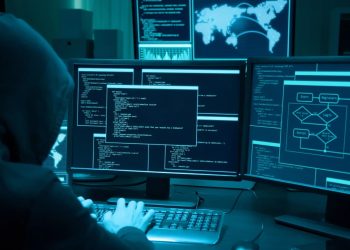 Hooded person working at a multi-monitor setup with glowing blue code and maps on the screens, creating a cybersecurity scene.