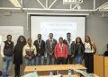 Group of nine diverse young people posing in a classroom in front of a projector screen that says 'For the People Hackathon', several wearing medals around their necks.