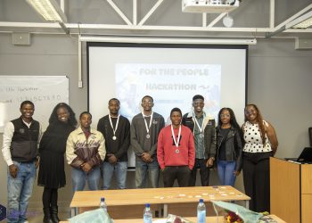 Group of nine diverse young people posing in a classroom in front of a projector screen that says 'For the People Hackathon', several wearing medals around their necks.