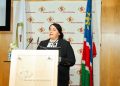 Woman in a black blazer delivers a speech at a wooden podium, Bank of Namibia backdrop behind her.