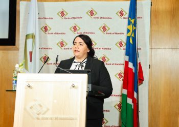 Woman in a black blazer delivers a speech at a wooden podium, Bank of Namibia backdrop behind her.