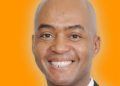 Head-and-shoulders portrait of a smiling bald man in a dark suit and white shirt against an orange background.