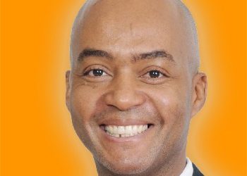 Head-and-shoulders portrait of a smiling bald man in a dark suit and white shirt against an orange background.