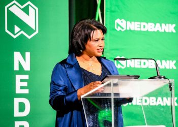 Woman in a navy blazer delivering a talk at a transparent podium with a tablet, a Nedbank green backdrop behind her.