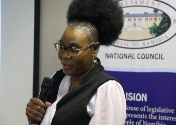 Woman with glasses speaking into a handheld microphone at a National Council event, banner in the background.