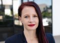 Portrait of a woman with dark red hair and red lipstick, wearing a navy blazer, outdoors in front of a modern glass building.