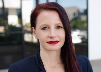 Portrait of a woman with dark red hair and red lipstick, wearing a navy blazer, outdoors in front of a modern glass building.