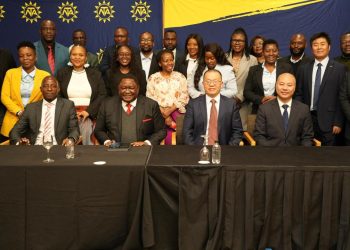 Group of diverse professionals posing for a formal group photo at a conference, seated at a long table with a black cloth and a blue backdrop with yellow logos behind them.