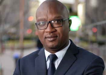 Close-up of a man in a navy suit and tie wearing black-rimmed glasses, outdoors with a blurred background.