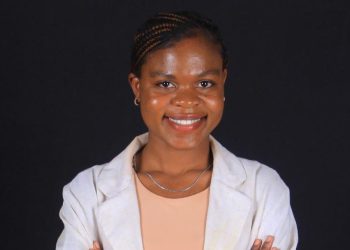 Smiling woman with braided hair wearing a light blazer and peach top against a black studio background.
