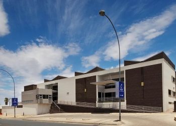 Exterior view of a modern white and dark-brick campus building with stairs, ramp access, and blue NQA banners under a blue sky. This shows the main entrance area.