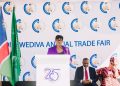 Woman in a purple blazer speaks at a white podium during the Wediva Annual Trade Fair, with flags and a logo-filled backdrop behind her.