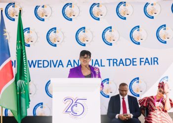 Woman in a purple blazer speaks at a white podium during the Wediva Annual Trade Fair, with flags and a logo-filled backdrop behind her.