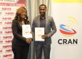 Two professionals in business attire shake hands while holding documents at a CRAN event, with banners in the background.