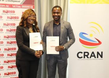 Two professionals in business attire shake hands while holding documents at a CRAN event, with banners in the background.