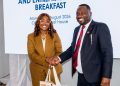 Two colleagues shake hands at a breakfast event; woman wears a tan suit with glasses and pearls, man wears a navy suit with a red tie, both smiling.