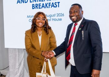 Two colleagues shake hands at a breakfast event; woman wears a tan suit with glasses and pearls, man wears a navy suit with a red tie, both smiling.