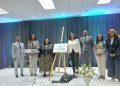 Group of eight professionals in business attire on a stage beside an easel with a DigiNam sign, posing for a photo at a formal event.